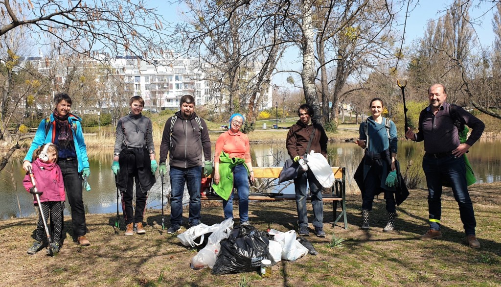 Einige Menschen stehen in einem Park vor einem Gewässer und haben gefüllte Mistsäcke vor sich stehen.