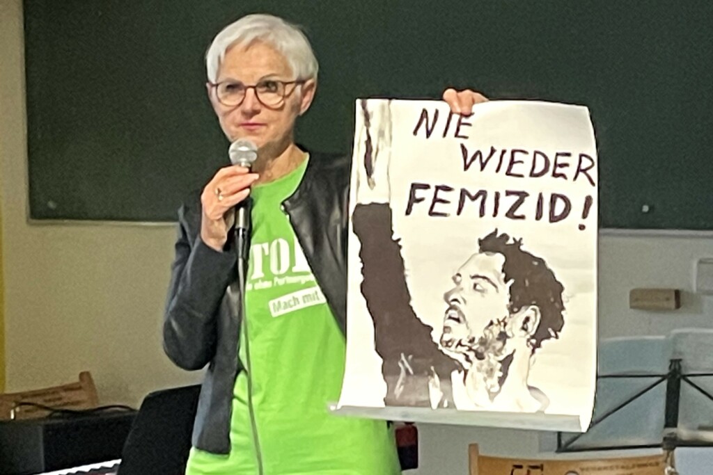 Frau spricht ins Mikrofon und hält ein Plakat in die Höhe mit dem Text "Nie wieder Femizid"