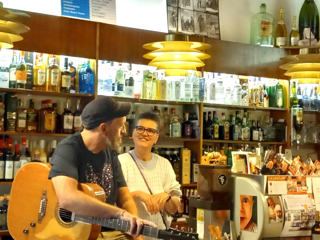 Ein Mann mit Gitrre und eine Frau hinter einer Schank mit vielen Flaschen