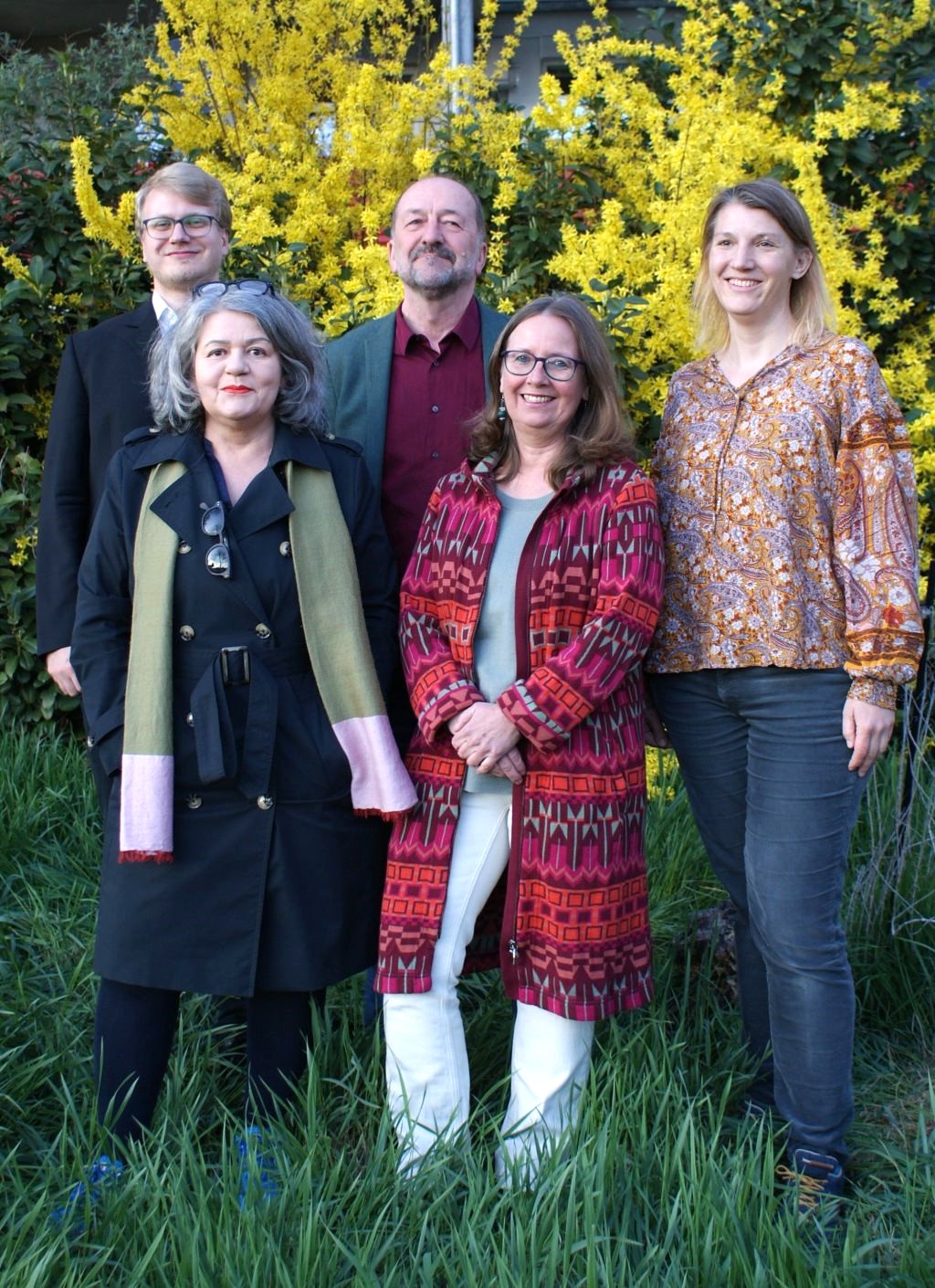 Fünf Personen stehen in einer Wiese vor einem blühenden Strauch und lächeln.