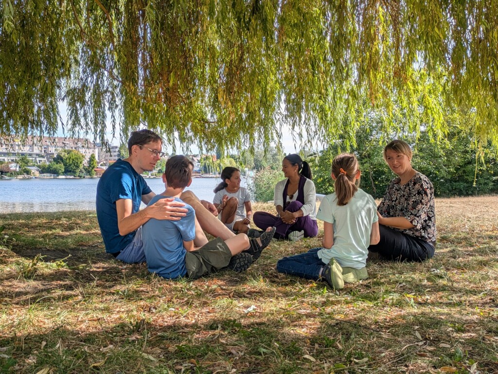 Erwachsene und Kinder siten im Kreis in einer Wiese und reden miteinander