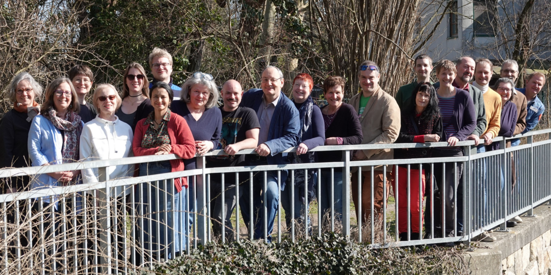 21 Menschen stehen lächelnd an einem brückenartigen Geländer in der Natur